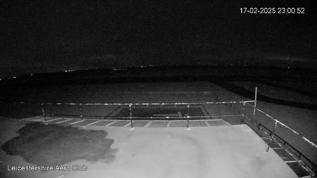 A dark scene showing an empty area with a railing in the foreground. The ground appears flat with parking lines visible. In the distance, faint lights can be seen, suggesting a landscape or horizon. The image is timestamped at 23:00 on February 17, 2025, and features a logo for the Leicestershire Aero Club.