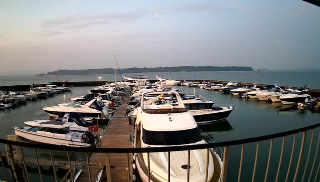 A panoramic view of a marina at dusk, featuring numerous boats of various sizes parked at a wooden dock. The water reflects the surrounding environment and is calm. In the background, a faint outline of land can be seen against the horizon, and the sky is gradually transitioning to twilight with light clouds. The scene is peaceful, with some light visible on the boats and the dock area.