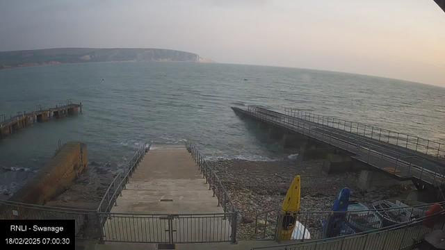 A coastal scene at dawn shows a beach with a boat ramp leading down to the water. To the left, a sturdy stone pier extends into the sea, and several boats are lined up along the shore. Yellow and blue kayaks are stored near the ramp. The water is calm, and the distant cliffs provide a scenic backdrop. The sky is light with soft colors, indicating early morning.