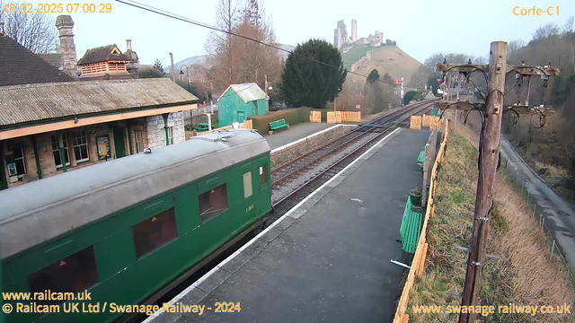 A green train carriage is positioned at a railway station platform, with a stone building partially visible behind it. To the left, there are green benches and a wooden fence enclosing some shrubs. In the background, a hill rises with the remains of a castle at the top. The sky is clear and light, suggesting an early morning. There is a telephone pole on the right with several wires extending from it.