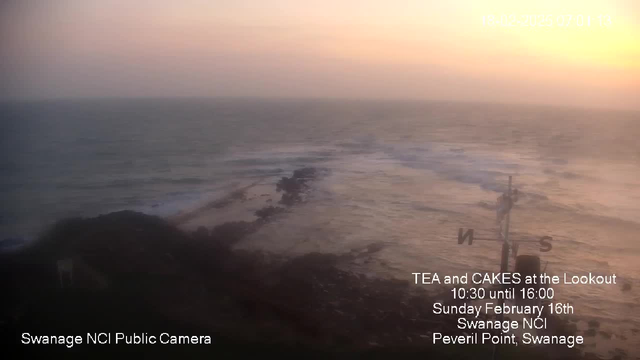 A coastal view at sunrise is depicted, showcasing gentle waves lapping against rocky shorelines. The sky transitions from soft pink to light orange as the sun rises. A weather vane stands in the foreground, indicating the North direction. Text in the bottom left corner notes it is a public camera from Swanage NCI, while additional text promotes a tea and cake event at a lookout point on February 16th. The overall atmosphere is calm and serene, suggesting a tranquil morning by the sea.