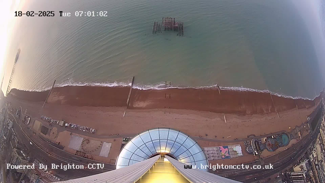 An aerial view of a beach with a long, sandy shoreline. In the foreground, a circular glass structure is visible, possibly part of a tower or observation deck. Below, the beach is dotted with amusement rides and structures. The water is calm, with gentle waves lapping at the shore. In the distance, a pier extends into the sea, while remnants of a structure can be seen submerged in the water. The scene is set in early morning light, with a soft glow illuminating the area. The time and date overlay indicates it is 7:01 AM on February 18, 2025.