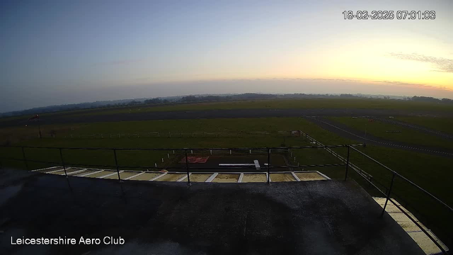 A view from a webcam located at Leicester Aeroclub during early morning. The sky is light blue with some clouds as the sun begins to rise. In the foreground, there is a railing surrounding an observation area, revealing a grassy airfield below. A long runway is visible, stretching into the distance with white markings. The landscape is flat and green with a few trees in the background, and the setting suggests a peaceful, quiet morning atmosphere. The timestamp at the top right indicates the date and time of the image.