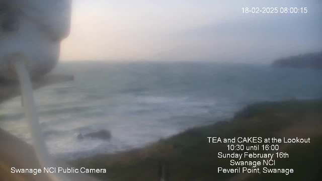 A blurred image depicting a coastal view with rolling waves. The ocean is visible in the background under a hazy sky. In the foreground, there is a slight obstruction, possibly part of the camera setup, on the left side. The bottom right corner includes text with event details: "TEA and CAKES at the Lookout, 10:30 until 16:00, Sunday February 16th, Swanage NCI, Peveril Point, Swanage."