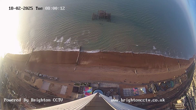 A high-angle view of a beach scene shows golden sand bordered by gentle waves from the sea. In the background, a dilapidated wooden pier stands partially submerged in the water. The foreground features a promenade with colorful buildings and attractions. The sky is bright, indicating early morning light. The image has a timestamp indicating the date as February 18, 2025, and the time as 08:00:12. The bottom left corner includes a logo that reads "Powered By Brighton CCTV" and a website URL.