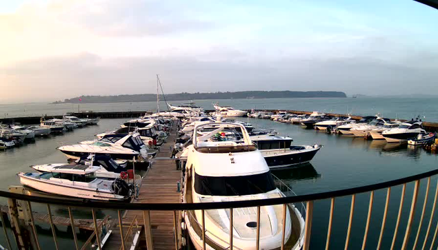 A wide view of a marina filled with various boats moored in the water. The scene includes several white and blue yachts lined up along a dock. In the background, there is a calm body of water with a distant landmass visible. The sky appears cloudy with soft light, suggesting either early morning or late afternoon. A railing in the foreground adds depth to the image.