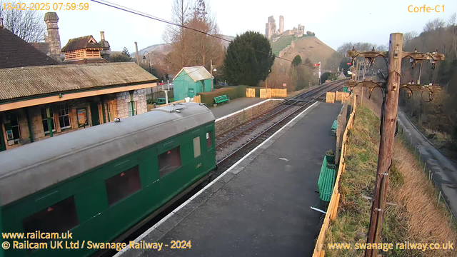 A green train carriage is partially visible on a platform at Corfe Castle railway station. The station building features a sloped roof and has large windows. There are green benches on the platform and a wooden fence in the background. A hill rises in the distance, topped by a castle ruin. The sky is bright with early morning light, and there are trees nearby. A wooden utility pole with wires is positioned on the right side of the image.