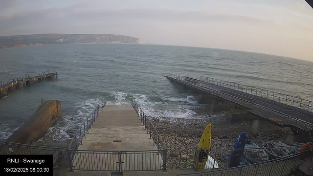 A view of the sea from a webcam, featuring a set of steps leading down to the water. On the left, there is a stone pier extending into the ocean, with railings along the edges. To the right, another pier stretches out towards the horizon. The water appears choppy with waves gently lapping at the shore, and in the foreground are several colorful kayaks stored near the steps. The sky is light with soft clouds, indicating early morning. The rocky beach is visible along the bottom of the steps.