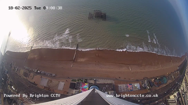 A bird's-eye view of a beachfront scene taken from a high vantage point. The image shows a sandy beach stretching along the coastline, with gentle waves lapping at the shore. An old, partially submerged pier is visible in the water. Amusement rides and colorful structures are located along the boardwalk, and several parked vehicles are discernible along the road adjacent to the beach. The sky is bright and clear, indicating daytime. Text in the corner displays the date and time, along with a logo indicating it is powered by Brighton CCTV.