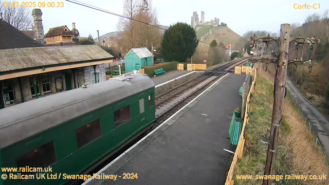 A view of a train station with a green train on the left side. The station building is made of stone, with a pitched roof and various windows. To the right, there are two railway tracks parallel to the platform. Behind the station, a hill features ruins of a castle, with trees and sparse vegetation surrounding it. A green shed is visible, and there are wooden benches and a fence along the platform. The scene is set during daytime with a clear sky.