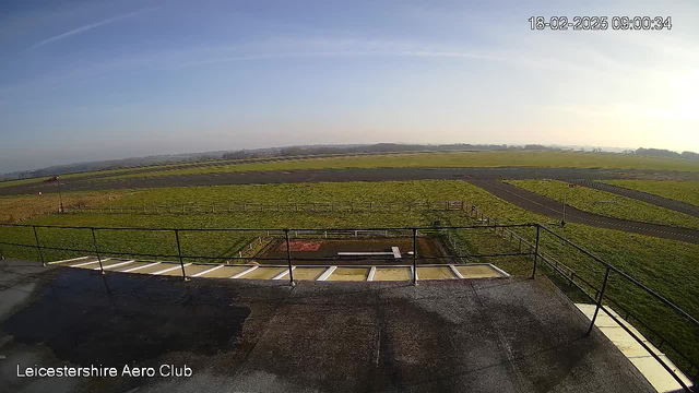 A wide view from an elevated position at Leicestershire Aero Club shows a grassy airfield stretching into the distance. The ground is mostly flat with patches of grass and some areas featuring a fence. There is a small structure or platform in the foreground, with a railing running along the edge. In the background, a clear blue sky is seen, indicating good weather, and additional land can be observed beyond the airfield. The time and date displayed in the corner read 09:00:34 on February 18, 2025.