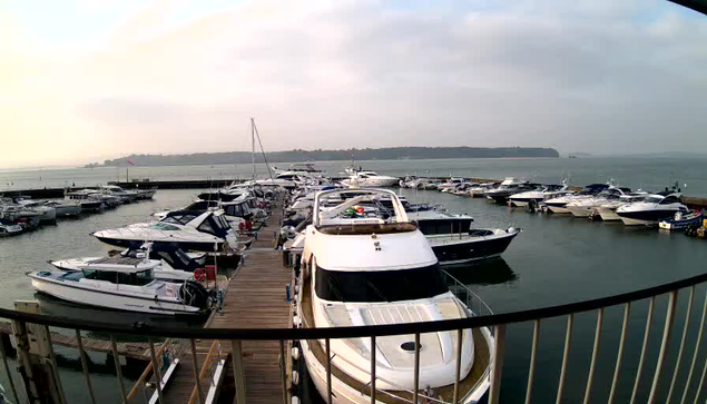 A view of a marina filled with various boats moored in the water. In the foreground, several yachts are visible, with some docked along a wooden walkway. The sky is overcast, and the water appears calm, reflecting the boats. In the background, a green landscape gradually rises, suggesting nearby land or hills. The scene conveys a peaceful, maritime atmosphere.