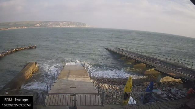 A view of the ocean from a pier, with steps leading down to the water. The sea shows gentle waves, and a rocky shore is visible at the bottom of the stairs. To the right, there are several boats, including a yellow one and a blue one, along with some equipment. In the background, a coastal landscape with slight hills and a cloudy sky can be seen.