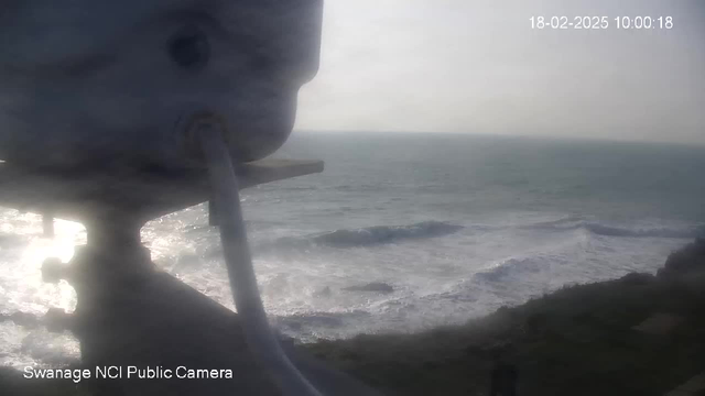 A blurred view from a webcam capturing the ocean waves rolling onto a shoreline. In the foreground, part of the webcam device is visible, with a white cable extending outward. The sky appears overcast, and the scene has a hazy quality, suggesting a calm but cloudy day at a coastal location. The bottom corner includes the text "Swanage NCI Public Camera" along with a timestamp.