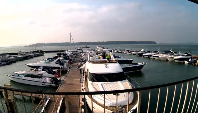 A marina scene with multiple boats docked in still water under a cloudy sky. In the foreground, there is a large white yacht moored at a wooden pier, surrounded by smaller vessels. The background features more boats, with a shoreline visible in the distance. The overall atmosphere is calm, suggesting a peaceful day at the marina.