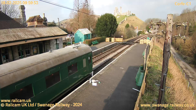 A view of a railway station with a green train parked on the platform. The scene includes a stone building with a sloped roof and several benches along the platform. In the background, a hill features a castle ruin. There are power poles to the right, and a green shed visible. The sky is partly cloudy, suggesting a clear day. The railway tracks extend to the horizon, while some decorative fencing can be seen near the entrance to the platform.