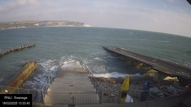 A coastline view featuring a serene sea in shades of blue. In the foreground, a concrete ramp leads down to the water, bordered by metal railings. To the right, a wooden jetty extends out over the waves, while a rocky shore is visible to the left. A yellow kayak and a blue storage container are placed nearby on the rocks. The sky above is partially cloudy.