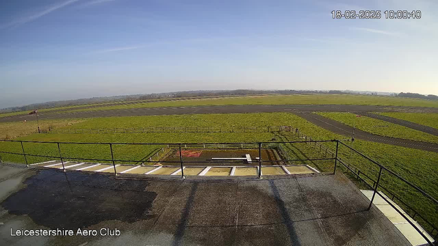 A wide view of an airfield featuring green grass and a large gray runway. In the foreground, there is a low railing and a concrete platform. The sky is clear with a few clouds, indicating a sunny day. In the distance, blurred green hills can be seen beyond the airfield. A light breeze can be inferred, as a flag is visible, gently fluttering in the wind.