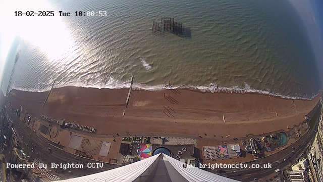 Aerial view of a sandy beach with light waves lapping at the shore. The beach is lined with a pier that has remnants of an old structure emerging from the water. In the foreground, colorful patterns on the ground suggest a carnival or amusement area. The beach is relatively empty, with a few people walking along the shore. The sky above is clear with minimal clouds, and sunlight reflects off the water's surface, creating a shimmering effect. The camera timestamp shows the date and time, and logos indicating the source of the image are present at the bottom.