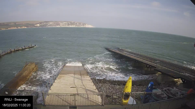 A coastal scene featuring a wooden pier extending into the bluish-green sea. The water has gentle waves. In the foreground, there is a wide concrete ramp leading down to the water, flanked by metal railings. To the right, there are several boats, including a yellow and a blue kayak, positioned on the rocky shore. In the background, cliffs rise against a light blue sky with some scattered clouds. The overall atmosphere is serene and maritime.