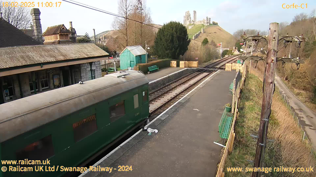 A train carriage in green is parked on a railway platform at Corfe Castle station. The platform features a low stone wall, wooden benches, and a green shed. In the background, a hill with the ruins of Corfe Castle is visible, surrounded by trees and open landscape. The sky is clear, indicating a sunny day. A wooden electricity pole stands to the right, with wires extending across the scene. The date and time displayed in the top left indicate it is 11:00 on February 18, 2025.