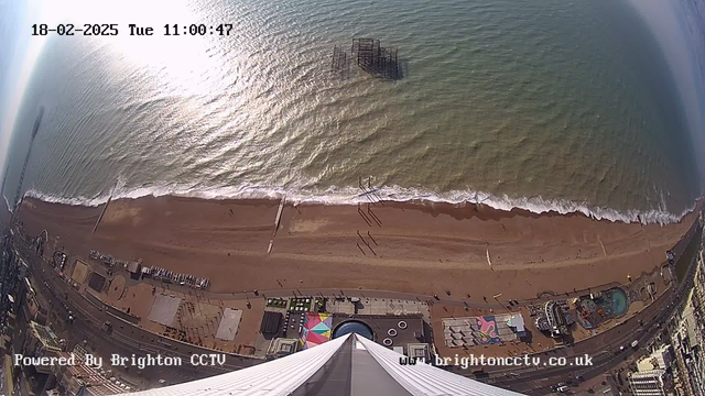 An aerial view of a beachfront. The image shows a sandy beach with gentle waves rolling in from the sea. There are people walking along the shore, and a pier structure is partially submerged in the water. In the foreground, various seaside attractions and structures, including a circular fountain and an amusement area, are visible. The sunlight reflects off the water, creating a shimmering effect. The date and time are displayed at the top of the image.