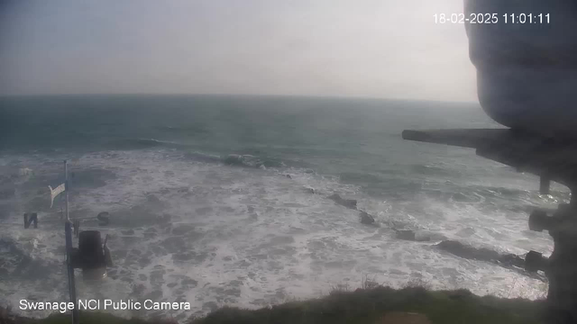 A coastal scene showing turbulent ocean waves crashing against rocks. The water appears choppy and frothy, with a slight mist in the air. In the foreground, there is a white flagpole with a flag and a structure partially visible on the right. The sky is overcast, hinting at possibly cloudy weather. The image is timestamped, indicating the date and time.