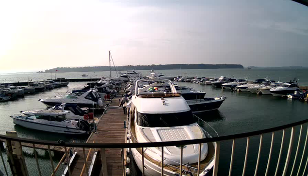 A marina scene with several boats docked at a wooden pier. The foreground features a large white yacht with a cabin and multiple smaller boats around it. In the background, a body of water stretches out towards a distant landmass under a cloudy sky. The overall atmosphere is calm and serene.