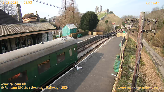 A green train sits on a railway platform with a wooden structure in the background. The scene is set under a clear sky, with a historic castle perched on a hilltop in the distance. The platform features several green benches and a small blue shed. Railway tracks run alongside the platform, leading off into the distance. There are bare trees and shrubs nearby, creating a natural landscape around the station.