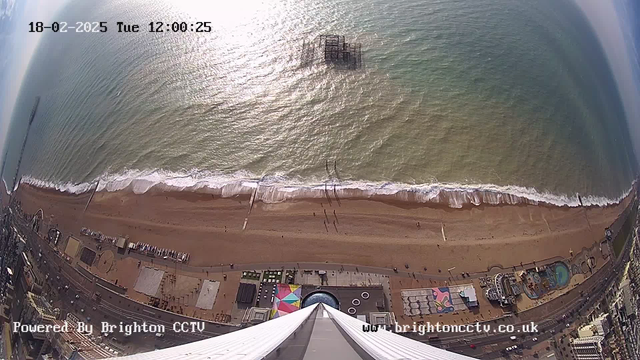 A view from a high vantage point overlooking a beach. The shoreline curves along the bottom of the image, with golden sand and gently rolling waves. A few people can be seen walking along the beach, with minimal activity. In the water, an old pier structure rises above the waves, partially submerged. The shoreline is bordered by a promenade with colorful beach huts or structures visible. The sky is mostly clear with some clouds, and the sun reflects off the water's surface, creating glimmers. An additional structure, possibly a ride or amusement area, is visible at the bottom of the image, alongside other buildings.