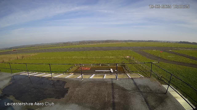 A clear blue sky with a few wispy clouds is visible above a wide, open field. In the foreground, there is a black railing on the terrace of a building, with a small puddle of water on the surface. Below the terrace, an airfield stretches out, featuring a grassy area on the left and a paved runway in the center. A red wind sock indicates wind direction, and a few scattered markers are visible on the grass. The landscape is peaceful, typical of an outdoor aviation space.