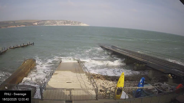 A coastal scene featuring a stairway leading down to a rocky shoreline with crashing waves. To the right, there are two kayaks, one yellow and one blue, secured on a deck. A wooden pier extends into the ocean on the left, with water visible all around and cliffs in the background. The sky is mostly clear with some clouds. The timestamp at the bottom indicates the date and time.