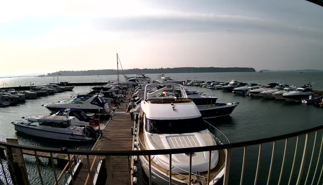 A view of a marina filled with numerous boats docked along a wooden pier. In the foreground, a large white yacht is moored at the edge of the dock. The water is calm, reflecting the cloudy sky above. Several smaller boats are also visible in the marina, with distant land and trees along the horizon.