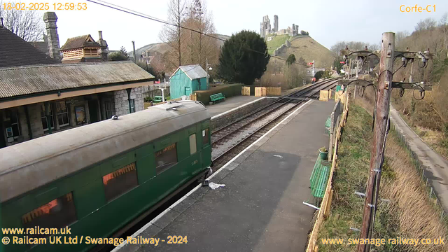 A green vintage train is parked at Corfe Castle railway station. In the background, there are trees and a hill with a historic castle ruin. The platform is lined with wooden benches and a green shed is visible. The sky is clear with some scattered clouds, and electrical poles are positioned along the side of the track. The atmosphere is serene and rural.