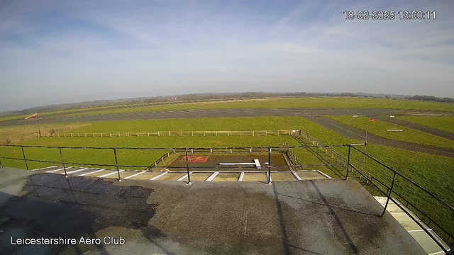 A view of a grassy airfield from a high vantage point, with a partially cloudy sky. The foreground shows a flat rooftop with metal railings and a small water area. In the distance, the airstrip is visible, lined with white markings, and a red windsock is on the left side. The landscape is mostly green with a few scattered trees and distant fields.