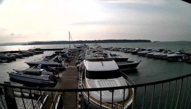 A waterfront scene featuring a marina filled with various boats docked in calm water. In the foreground, a large white yacht with a sleek design is prominently positioned on the left side. Several smaller boats are scattered along the dock, and the area is bordered by wooden walkways. The sky is overcast with soft, diffused light, and distant hills can be seen on the horizon.