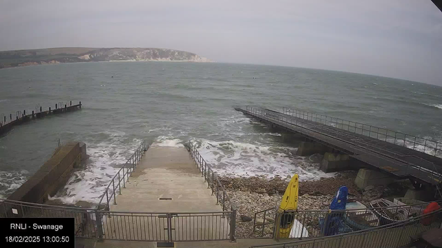 A view of a seaside area with a concrete staircase leading down to the water. On the left, there is a stone jetty extending into the waves. The ocean is choppy with white-capped waves, and the sky is overcast. To the right, there are two kayaks, one yellow and one blue, positioned on a gravel surface near the water. The scene captures the coastal environment with cliffs visible in the background.