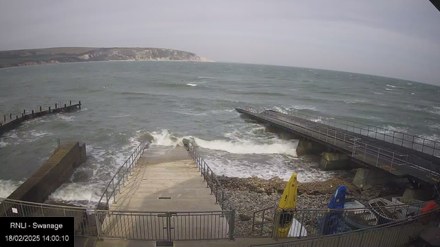 A view of a coastal area with rough waves crashing against the shoreline. In the foreground, there are concrete steps leading down to the water and a metal railing on one side. To the right, there are several small kayaks in yellow and blue, along with a few boats. In the background, a rocky coastline with hills is visible under a cloudy sky. The water appears choppy, indicating windy conditions.