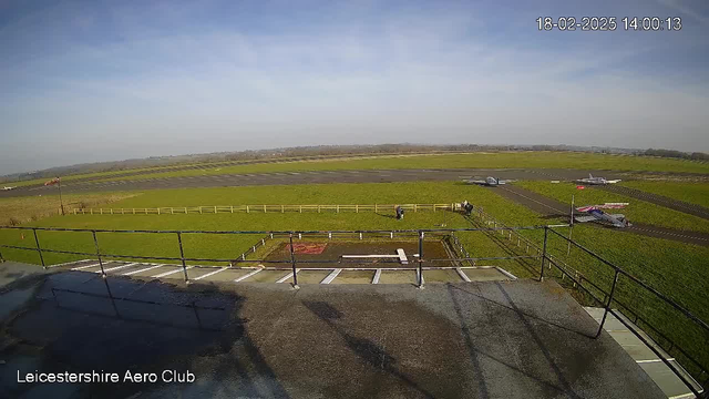 A view from a high vantage point at a grassy airfield. The foreground features a railing and part of the building structure. In the background, several small airplanes are parked on the tarmac, with a grassy area surrounding them. A few people can be seen walking on the ground near the planes. The sky is mostly clear with a few clouds and the date and time are displayed in the corner.