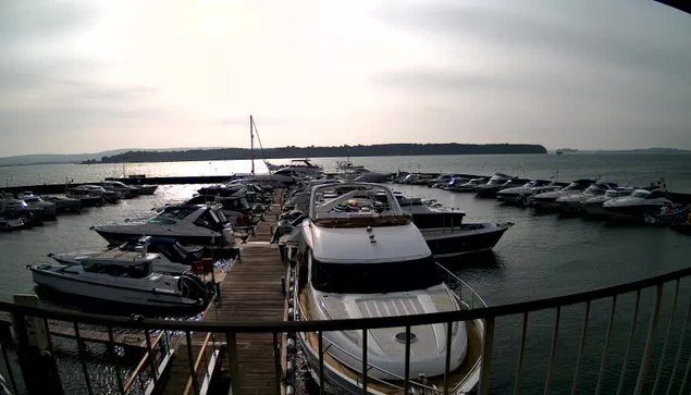 A view of a marina with numerous boats docked in the water. In the foreground, several boats are moored alongside a wooden pier. The sun reflects off the water, creating a shimmering effect. A distant shoreline is visible on the horizon, partially obscured by hazy clouds. The sky is overcast, contributing to a soft, diffused light over the scene.