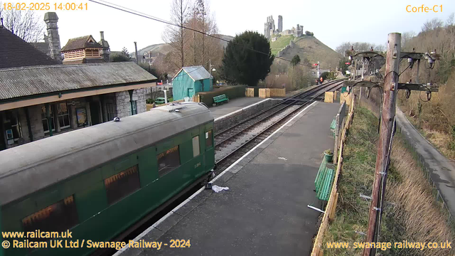 A view of a railway station with a green train parked alongside the platform. In the background, there is a hilly landscape featuring a castle ruins atop a hill. The station building is made of stone, with a sloped roof and several windows. Nearby, there are green benches and a small blue shed. There are wooden fences, power lines, and a distant road lined with trees. The sky is clear, indicating a sunny day.