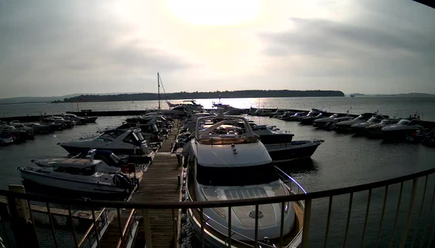 A waterfront scene featuring a marina filled with various boats docked along a wooden pier. The sun is low in the sky, casting a reflective light over the water. In the background, dark hills rise against a cloudy sky. Several boats are visible, including some larger ones in the foreground and smaller ones further back, creating a busy but serene atmosphere.