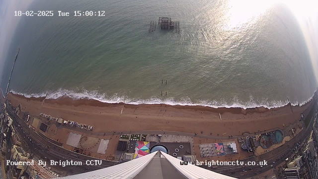 Aerial view of a sandy beach with waves gently rolling in. The shoreline has various structures, including a disused pier partially submerged in the water. There are colorful beach umbrellas and some buildings adjacent to the beach. The scene captures the ocean's surface reflecting sunlight, giving a bright appearance. The date and time display is visible in the corner of the image, along with the logo for Brighton CCTV.