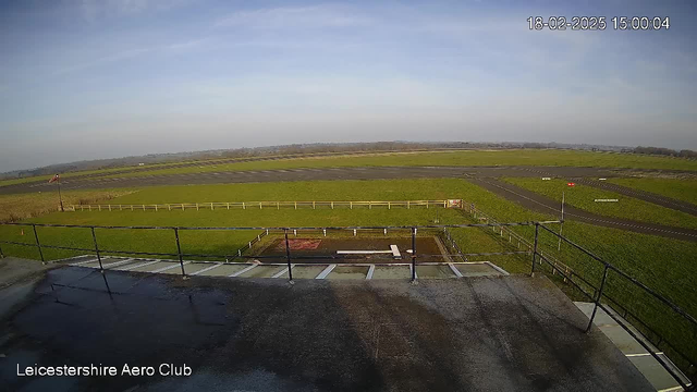 A wide view of an airfield on a clear day. The foreground shows a flat rooftop with a metal railing, partially covered in wet spots. Below, the airfield features green grass and a runway with a few markings. A white fence runs along part of the airfield, with a parking area visible. In the distance, there are low hills under a blue sky scattered with light clouds. The time and date are displayed in the corner.