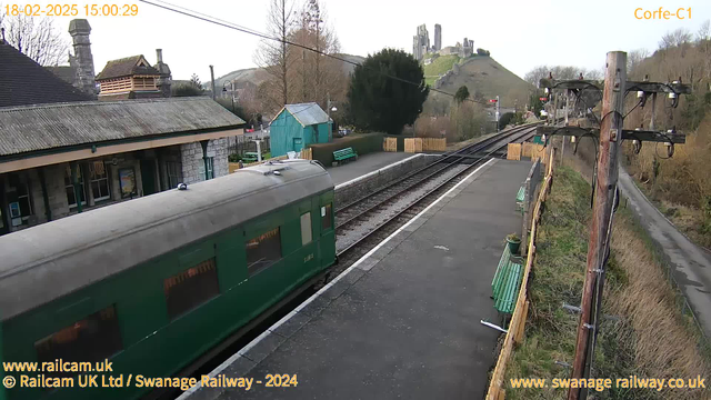 A green train is stationary at a railway station platform. The foreground shows part of the train and a paved area with a low stone wall. On the platform, there are green wooden benches. In the background, a quaint station building with a sloping roof and brick walls is visible. Beyond the station, a hill rises steeply, featuring ruins of a castle at the top. Trees and shrubs are scattered around the area, with a cloudy sky overhead. Power lines and a wooden pole stand along the edge of the platform.