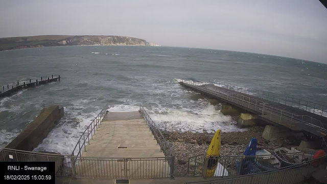 A rocky shoreline is shown with choppy waves approaching. In the foreground, there are concrete steps leading down to the water, flanked by railings. To the right, there is a wooden pier extending into the sea. Several colorful kayaks, yellow and blue, are positioned on the shore near the pier. In the background, there are cliffs and grassy hills covered with vegetation under a cloudy sky, stretching along the horizon.