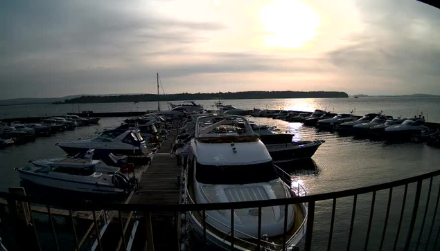 A marina with numerous boats docked on calm water. The scene is bathed in soft lighting with a cloudy sky, hinting at a sunset in the background. The water reflects the colors of the sky, creating a serene atmosphere. A wooden pier runs along the foreground where some boats are moored.