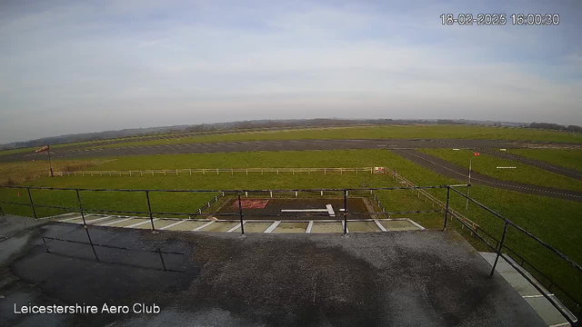 A panoramic view from a webcam overlooking a grassy airfield. The foreground shows a metal railing along a ledge. Below, there is a small area with a red patch and a white strip marking a landing zone. In the distance, the field stretches out with a few runway markings, and a light wind flag is visible on the left side. The sky is mostly clear with some clouds. A timestamp in the corner indicates the image was captured on February 18, 2025, at 16:00:30. The name "Leicestershire Aero Club" appears at the bottom.