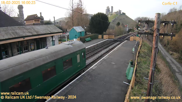 A green train car is positioned on the left side of the image, close to the edge of a train platform. In the background, a historic building with a stone facade is visible, along with a blue shed and several wooden benches placed along the platform. The railway tracks extend into the distance to the right, with a wooden fence and trees visible along the perimeter. On a hill in the background, there are the ruins of a castle. The sky is light and overcast. The timestamp at the top indicates the date and time of the image.