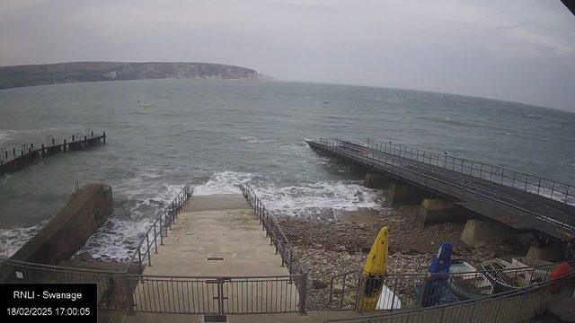 A coastal scene featuring a rocky shoreline with steps leading down to the sea. In the foreground, there are yellow and blue kayaks stored to the side. To the right, there is a wooden jetty extending into the water, while more rocky cliffs are visible in the distance. The sky is overcast, and gentle waves are rolling onto the shore.